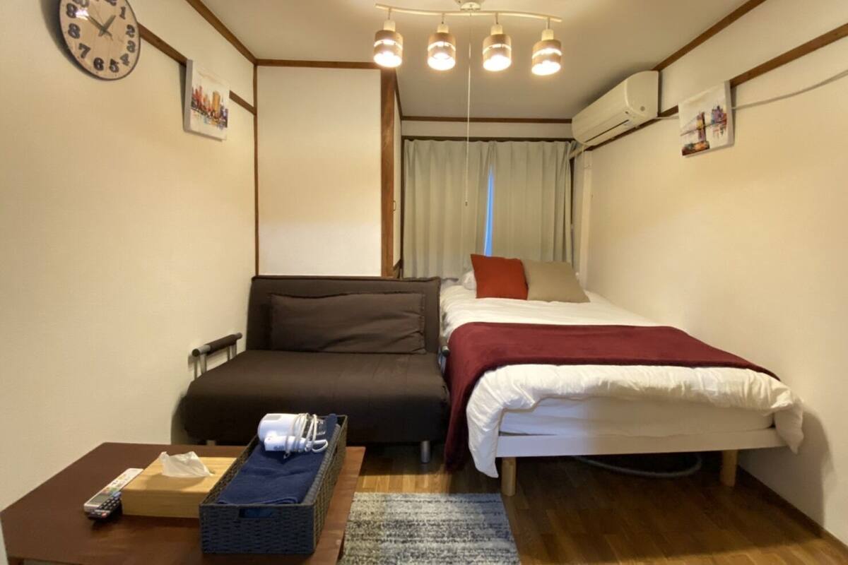 A compact hotel room with a bed, a sofa, a small table with a remote control, and a clock on the wall.