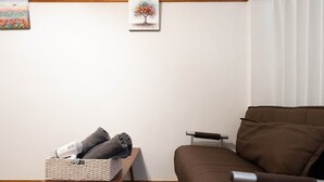 A living room with a brown sofa, a small table with a basket containing a hairdryer and towels, and two framed pictures on the wall. - Apartment Y Kawara Heights Namba (Osaka)