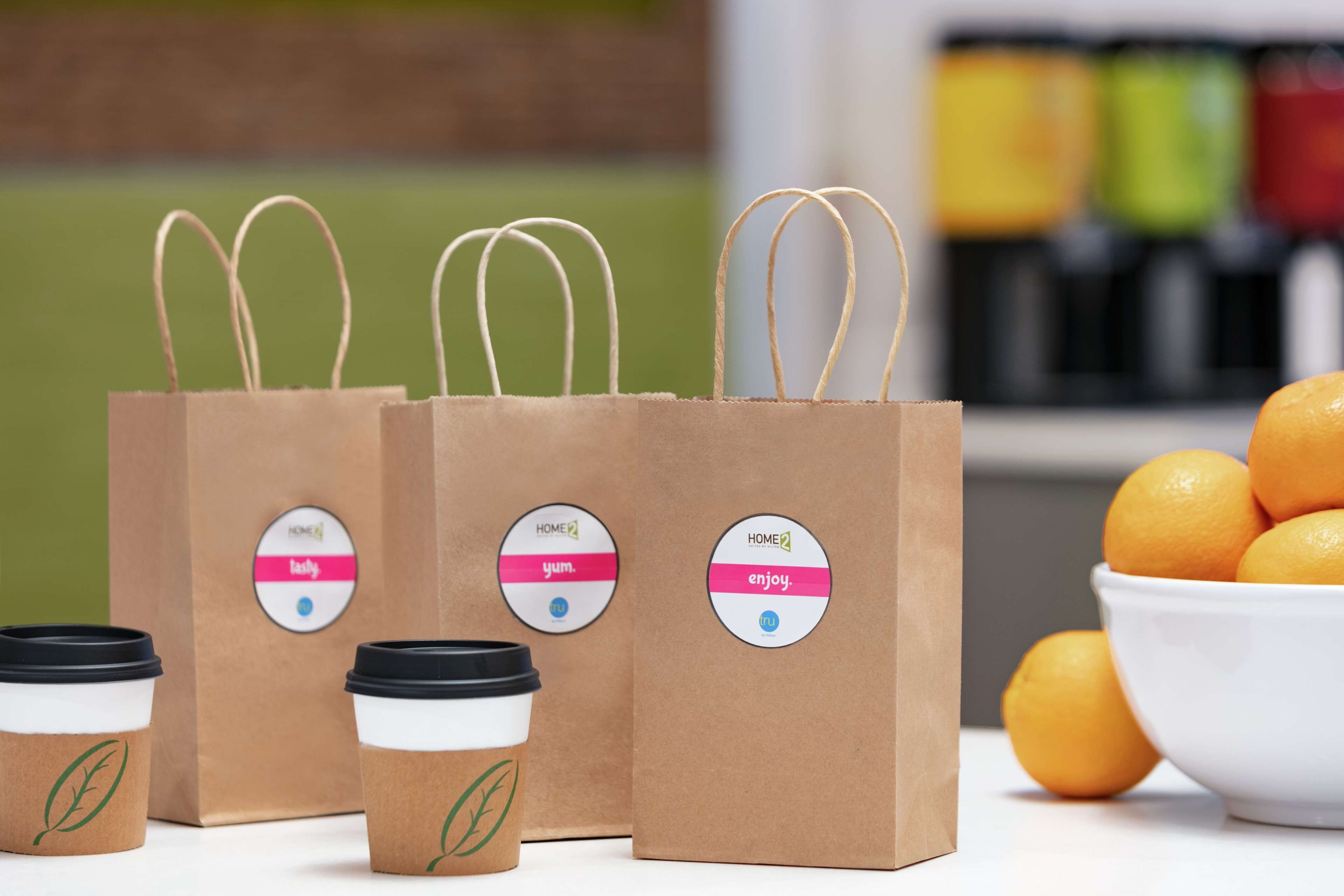 Three brown paper coffee bags with handles and lids, a bowl of oranges, and a blurred background with colorful containers.
