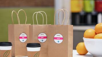 Three brown paper coffee bags with handles and lids, a bowl of oranges, and a blurred background with colorful containers.