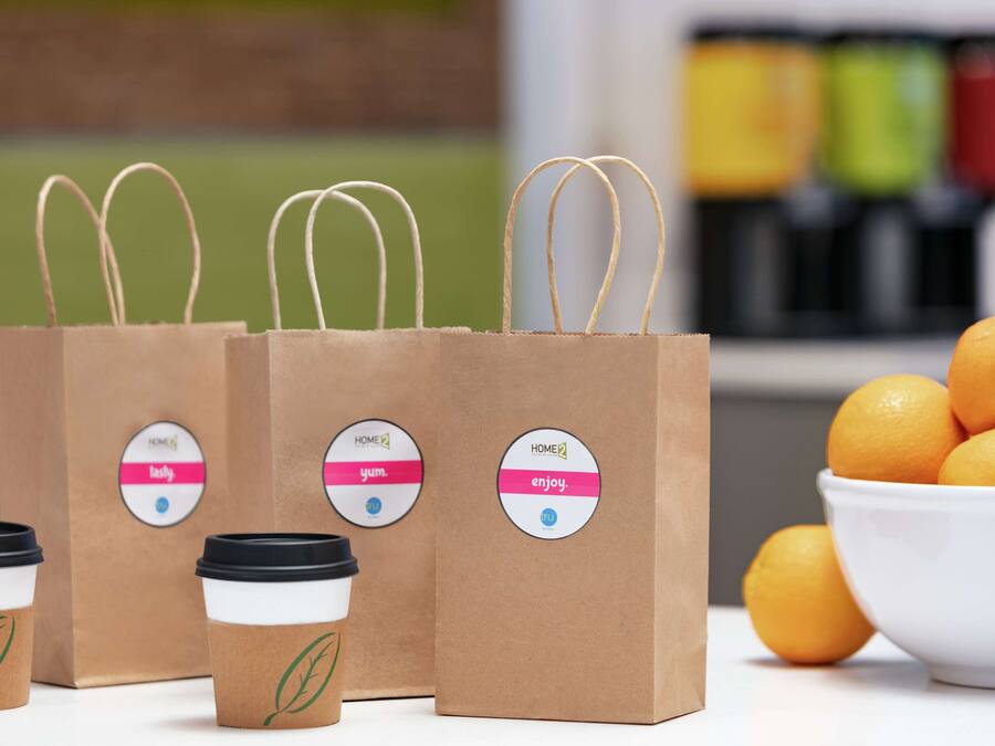 Three brown paper coffee bags with handles and lids, a bowl of oranges, and a blurred background with colorful containers.