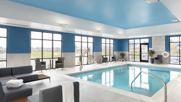 Un espace piscine intérieur moderne, avec un plafond bleu, de grandes fenêtres et une piscine équipée d’une échelle en métal.