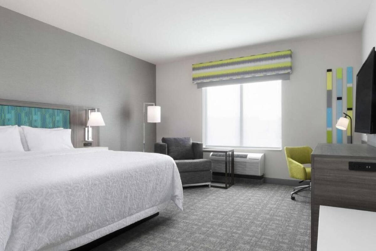 Habitación de hotel con cama, escritorio, silla, lámpara, ventana con persianas y televisor.