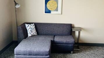 A gray sectional sofa with a patterned pillow, a small side table, and a floor lamp in a room with a plain wall and carpeted floor.