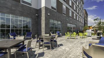 An outdoor patio with wicker furniture and a table, adjacent to a building with large windows and a grey facade.