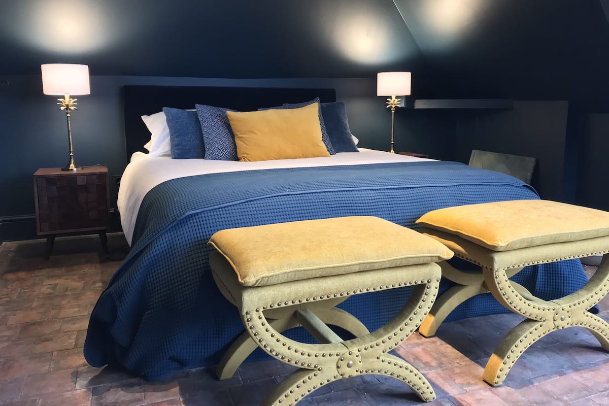 Quarto com uma cama grande, duas luminárias de cabeceira e dois banquinhos iguais.