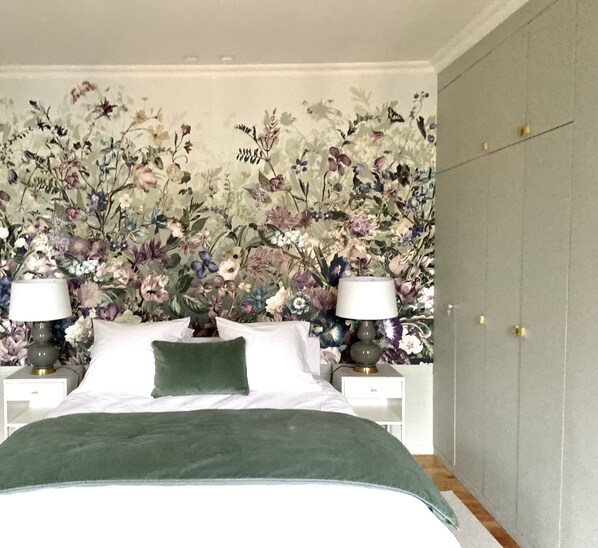 Un dormitorio con papel tapiz floral, una cama con ropa de cama verde, dos mesitas de noche con lámparas y un armario gris.