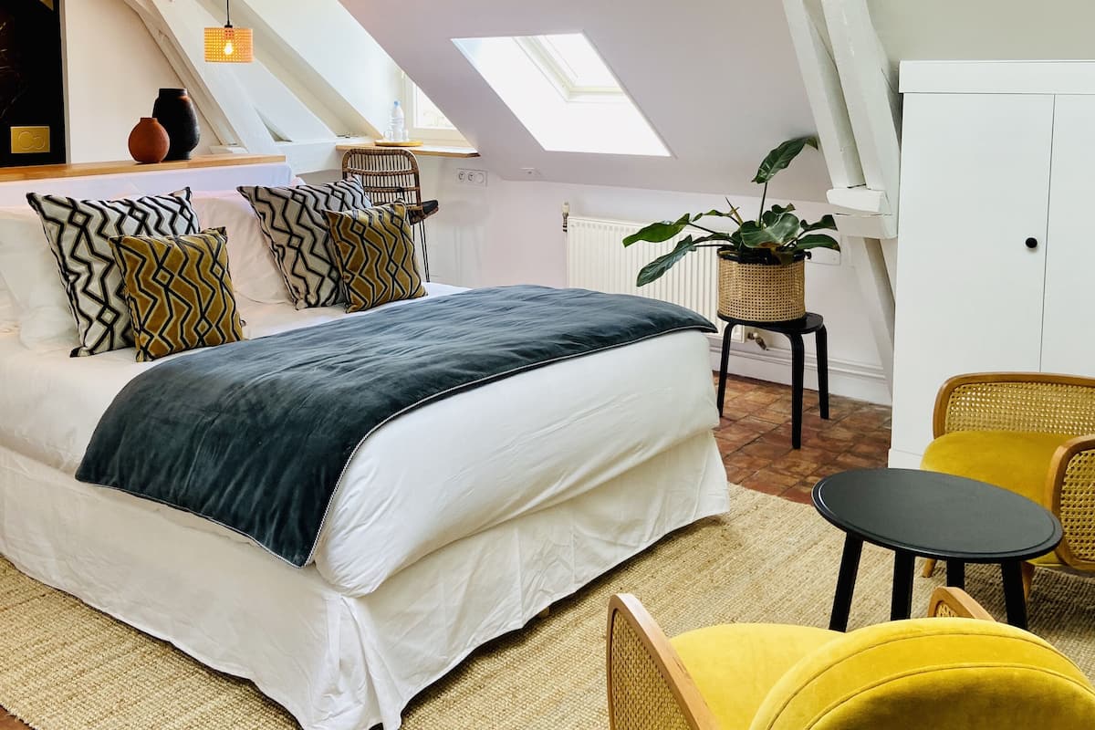 Quarto com cama, mesinha, cadeira, planta em vaso e claraboia.