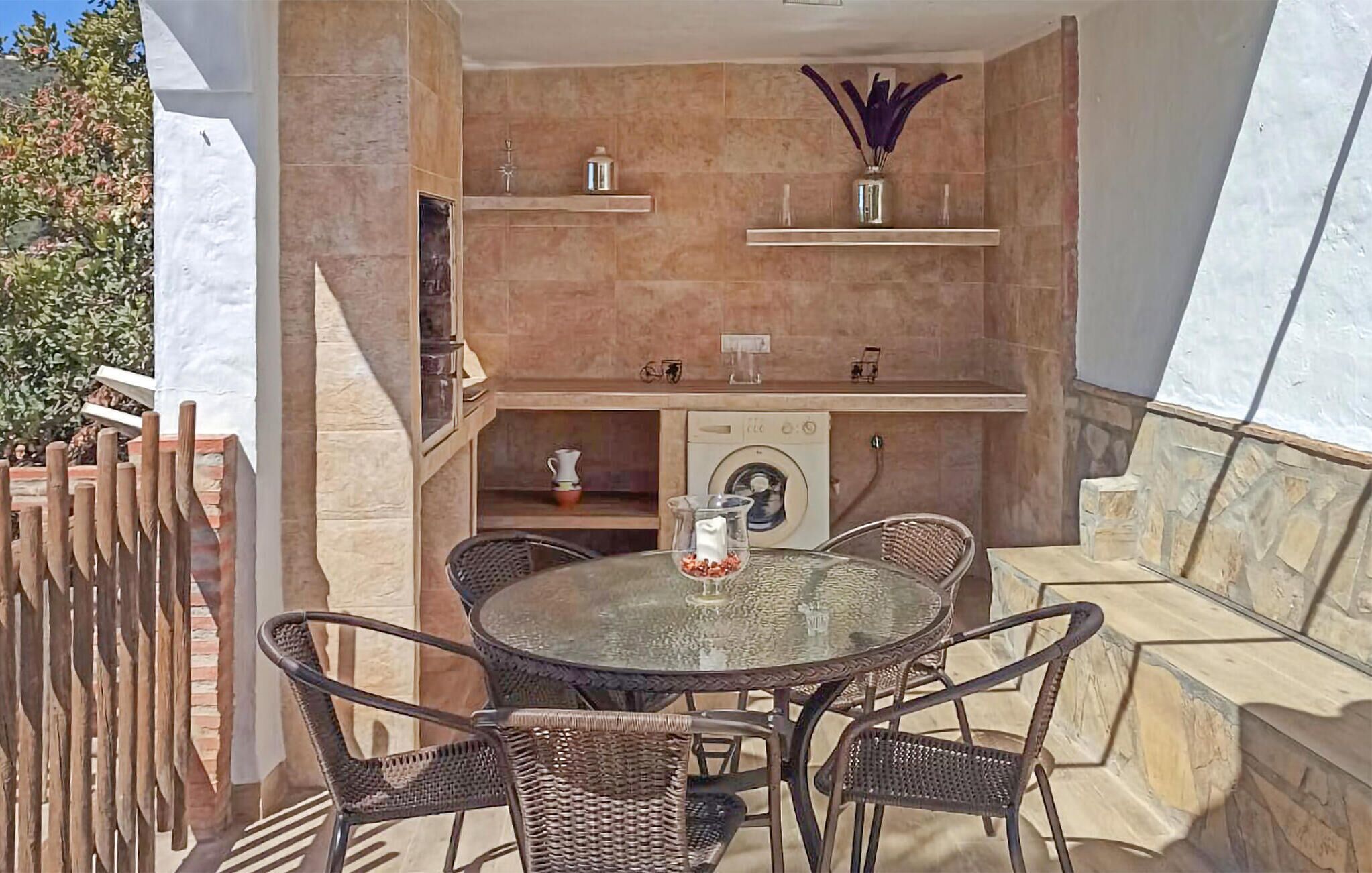 Une terrasse extérieure avec une table ronde en verre et des chaises en osier, un lave-linge et des étagères ornées d’objets décoratifs.