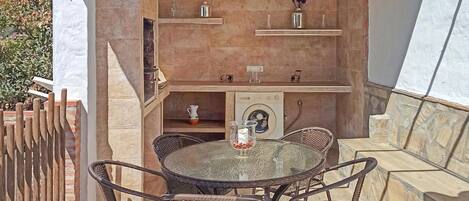Une terrasse extérieure avec une table ronde en verre et des chaises en osier, un lave-linge et des étagères ornées d’objets décoratifs.