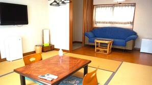 A room with a wooden table, chairs, a blue sofa, a television, and a window with curtains. - Resort Pension Turtle House (Shimoda)