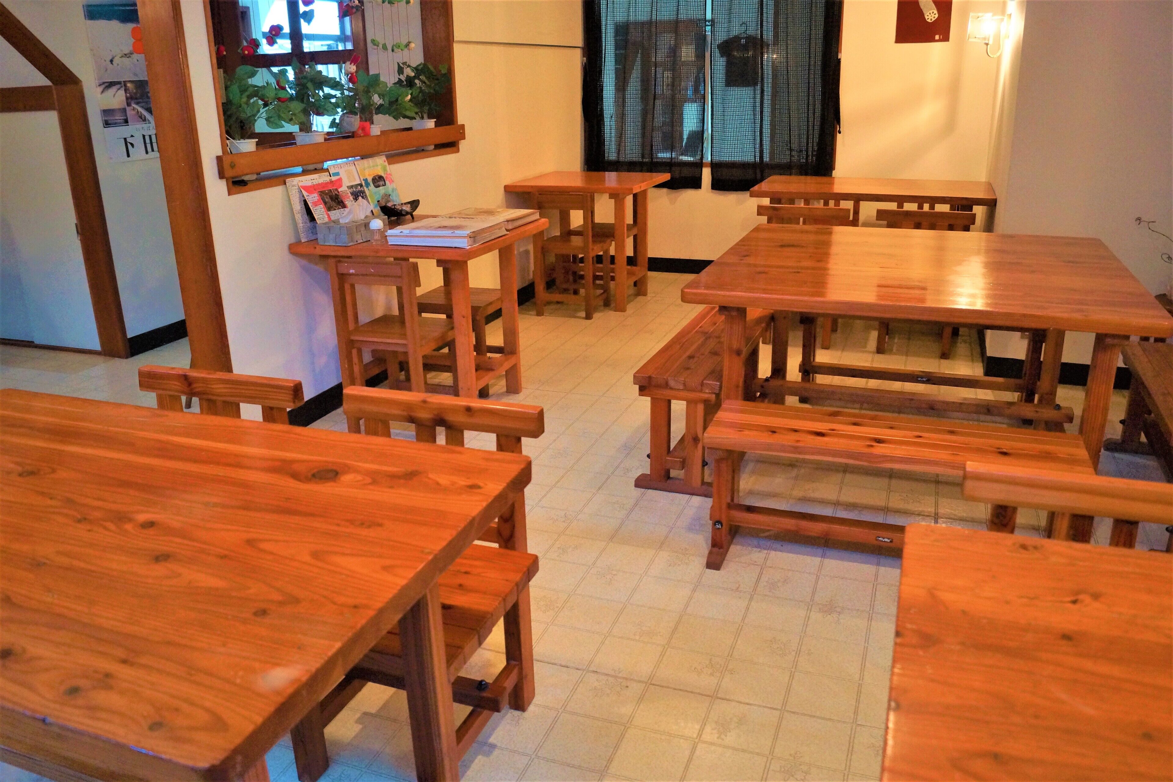 A dining area with wooden tables and benches, a window with a view, and a wall-mounted shelf with books and a potted plant.