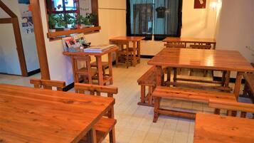 A dining area with wooden tables and benches, a window with a view, and a wall-mounted shelf with books and a potted plant.