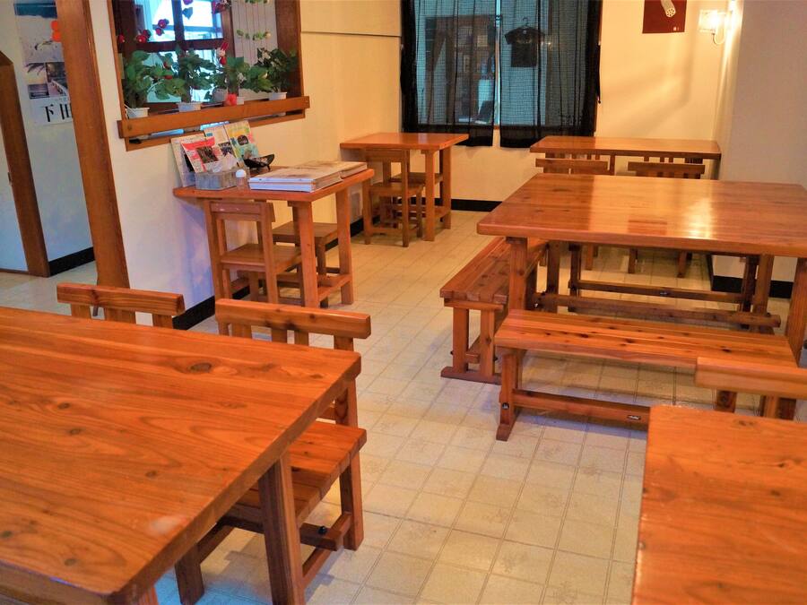 A dining area with wooden tables and benches, a window with a view, and a wall-mounted shelf with books and a potted plant.