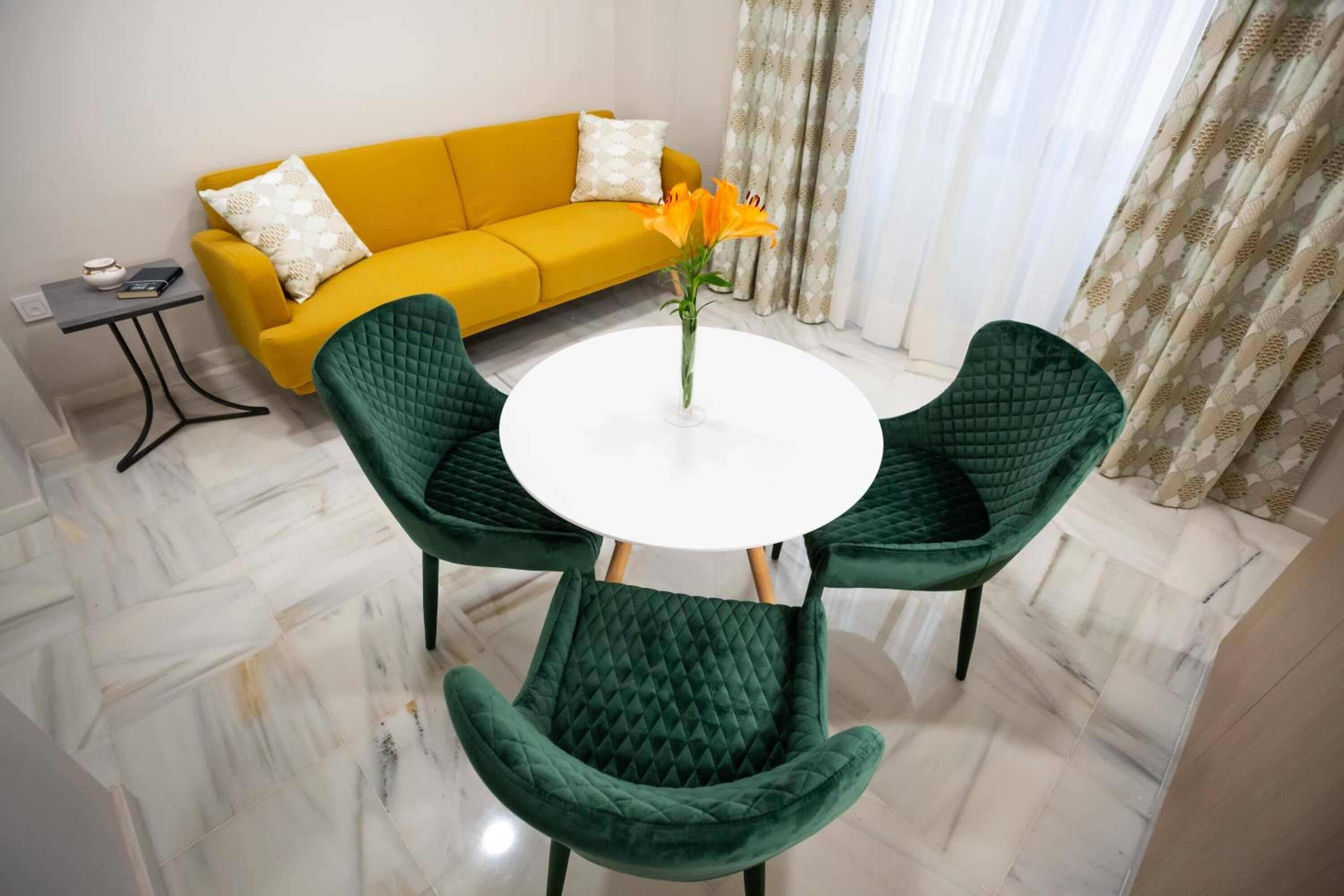 A room with a yellow sofa, a round table with chairs, and a vase with flowers.