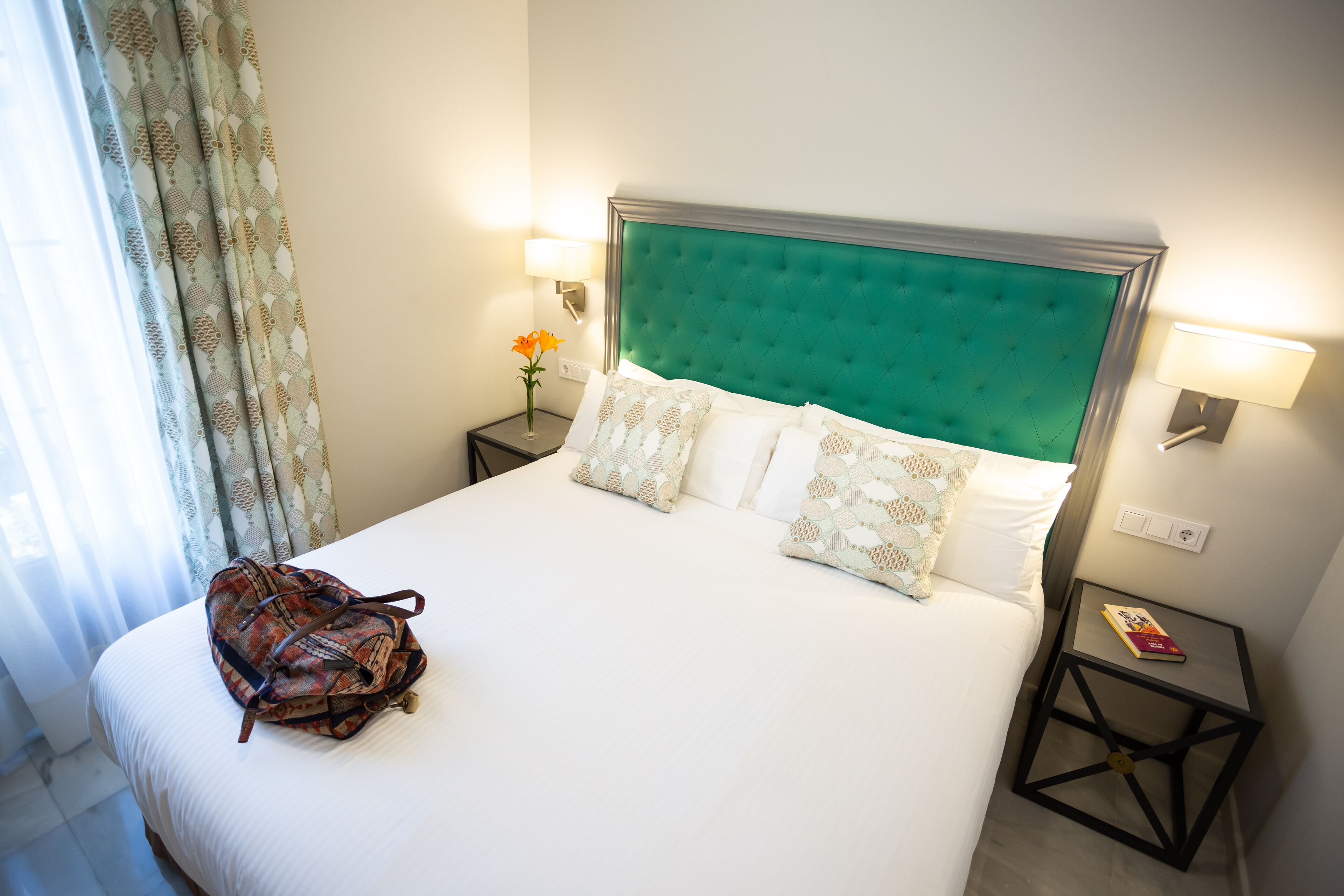 Un lit bien fait, avec une tête de lit verte, une literie blanche et des coussins décoratifs. Une petite table avec une lampe et un livre se trouve à côté du lit. Un sac à motifs est posé sur le lit.