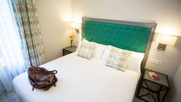 Cama bem arrumada com cabeceira verde, roupa de cama branca e almofadas decorativas. Pequena mesa com abajur e um livro ao lado da cama. Uma bolsa estampada está colocada sobre a cama.