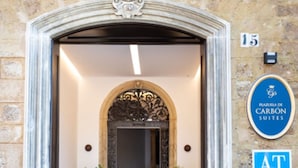 A hotel entrance with a stone facade, a decorative archway, a staircase leading inside, and a sign indicating the hotel name and address. - Plazuela del Carbón Suites (Cádiz)
