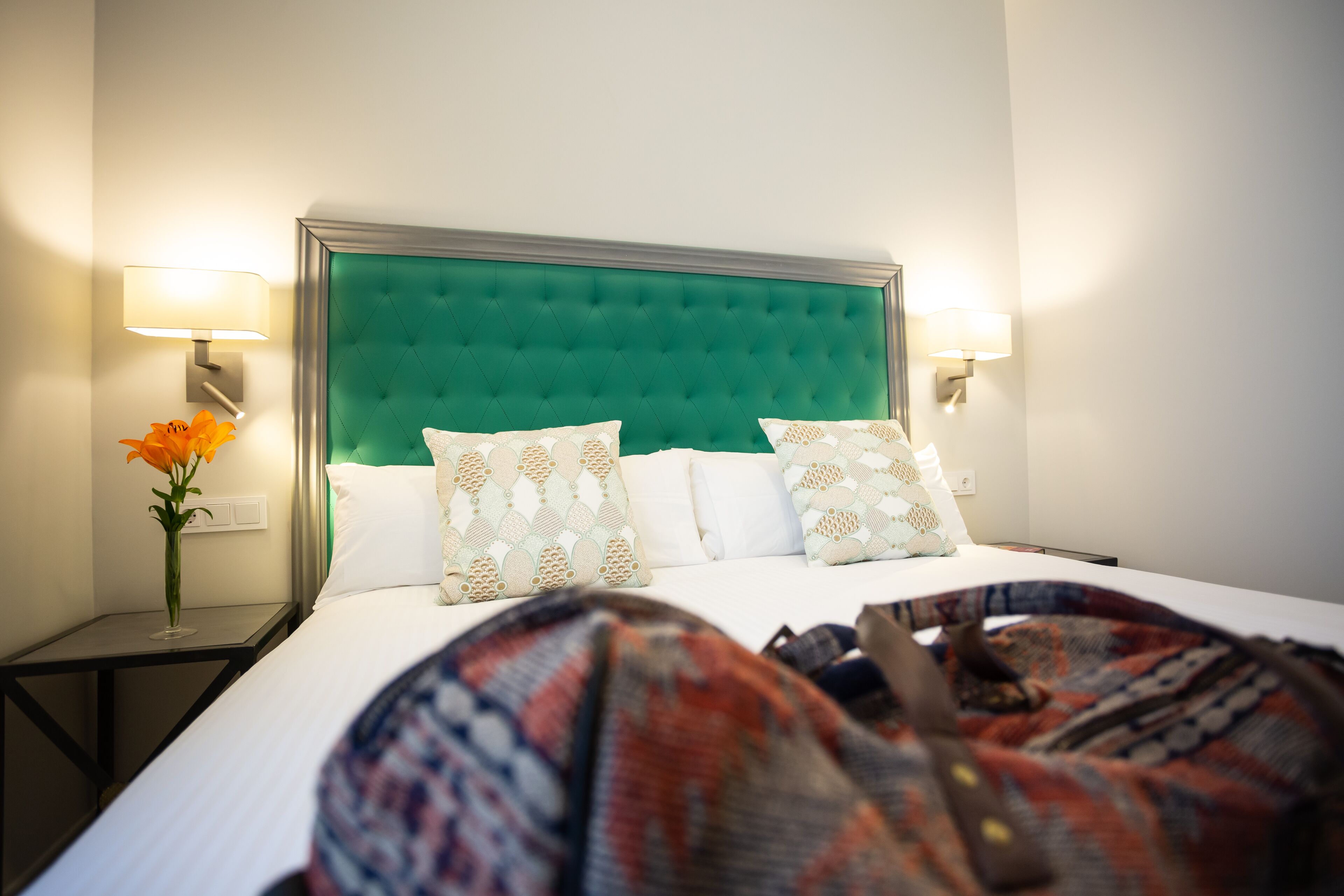 Habitación de hotel con una tapicería verde en la cabecera, ropa de cama blanca, almohadas estampadas y dos lámparas de noche.