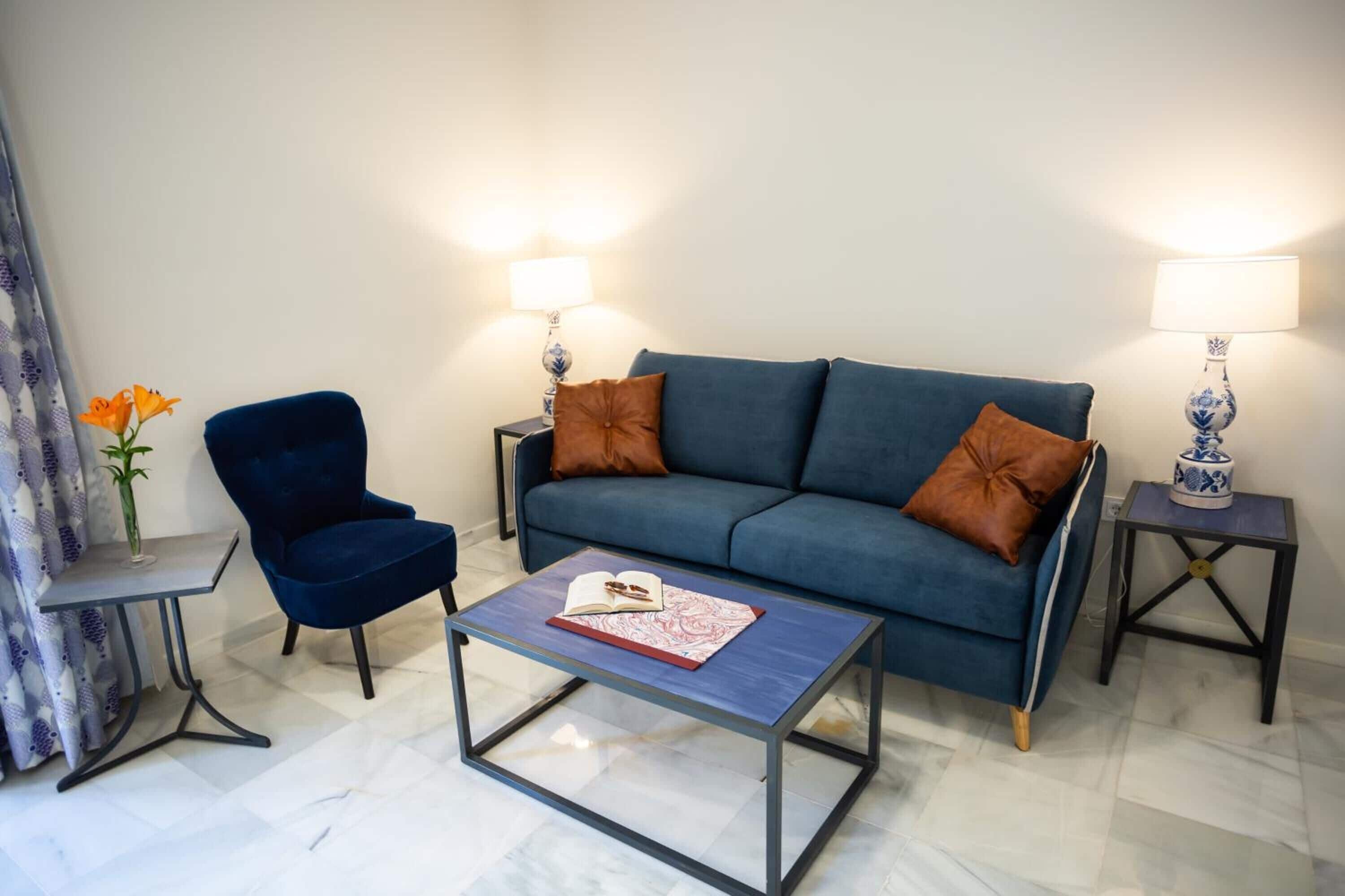 A living room with a blue sofa, two side tables with lamps, a blue chair, and a coffee table with a book and glasses.