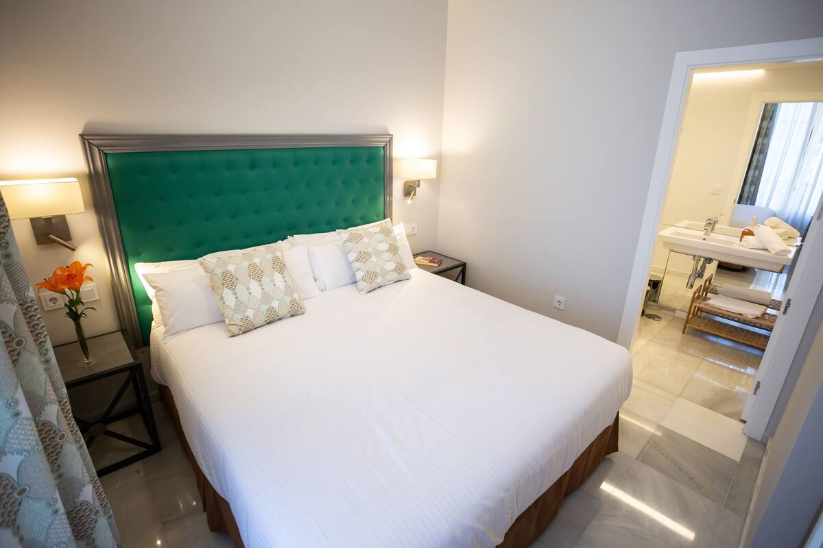 Cama bem arrumada com cabeceira verde e roupa de cama branca, mesa de cabeceira com luminária, e vista para um banheiro com pia e espelho.
