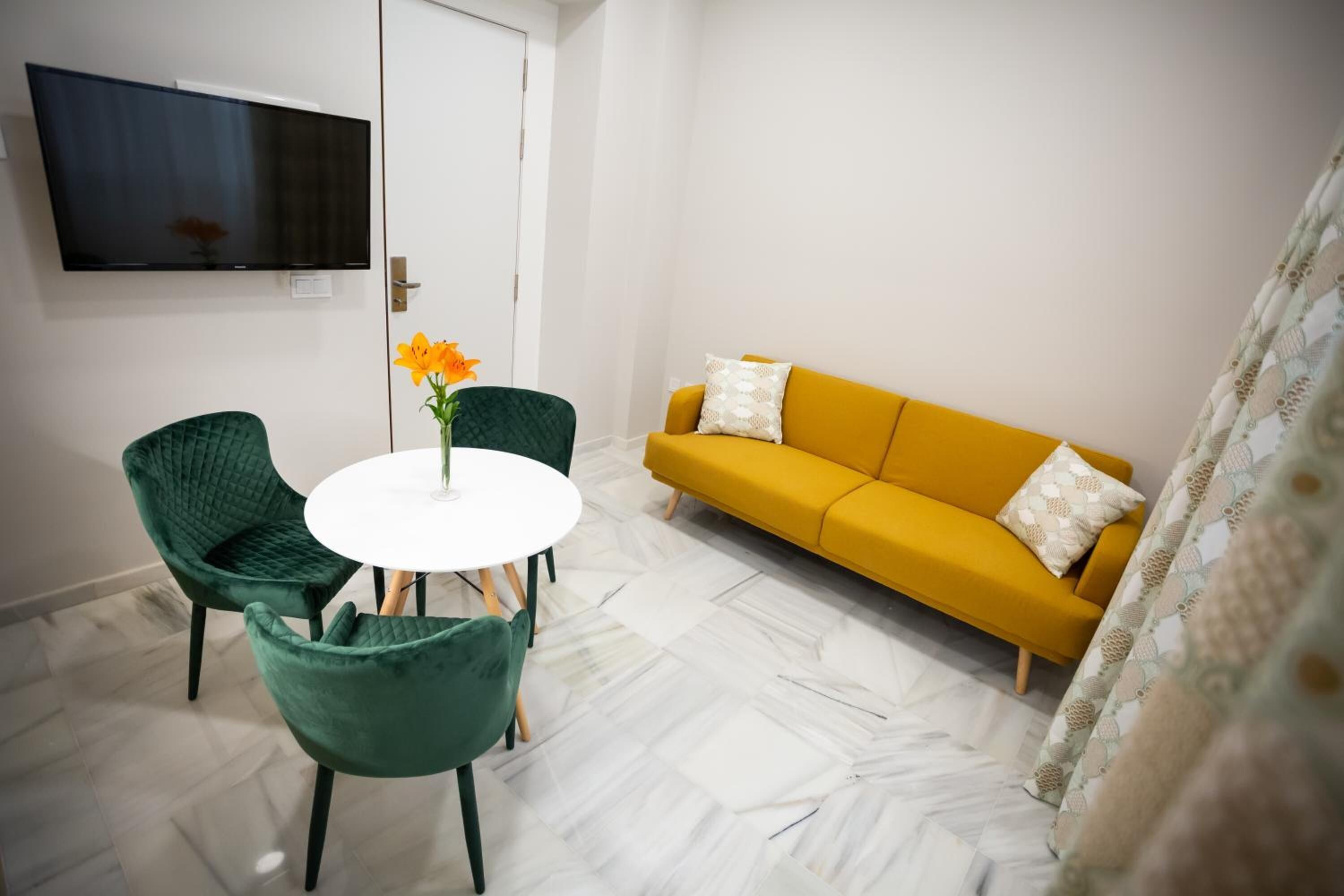 Een kamer met een witte ronde tafel, twee groene stoelen, een geel sofa en een flatscreen televisie aan de muur.