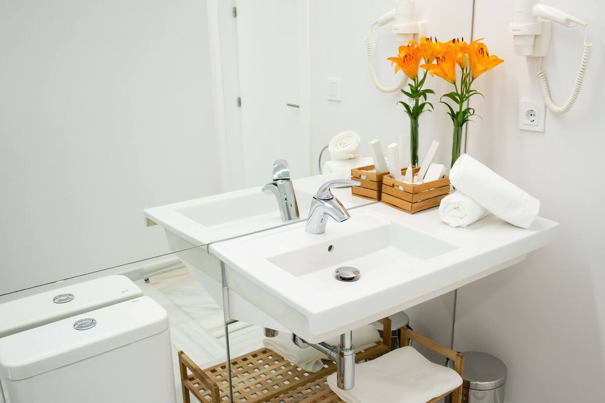 Um banheiro moderno com pia branca, duas espelhos e um vaso sanitário. Há flores laranja na pia, uma cesta de madeira e uma lixeira.