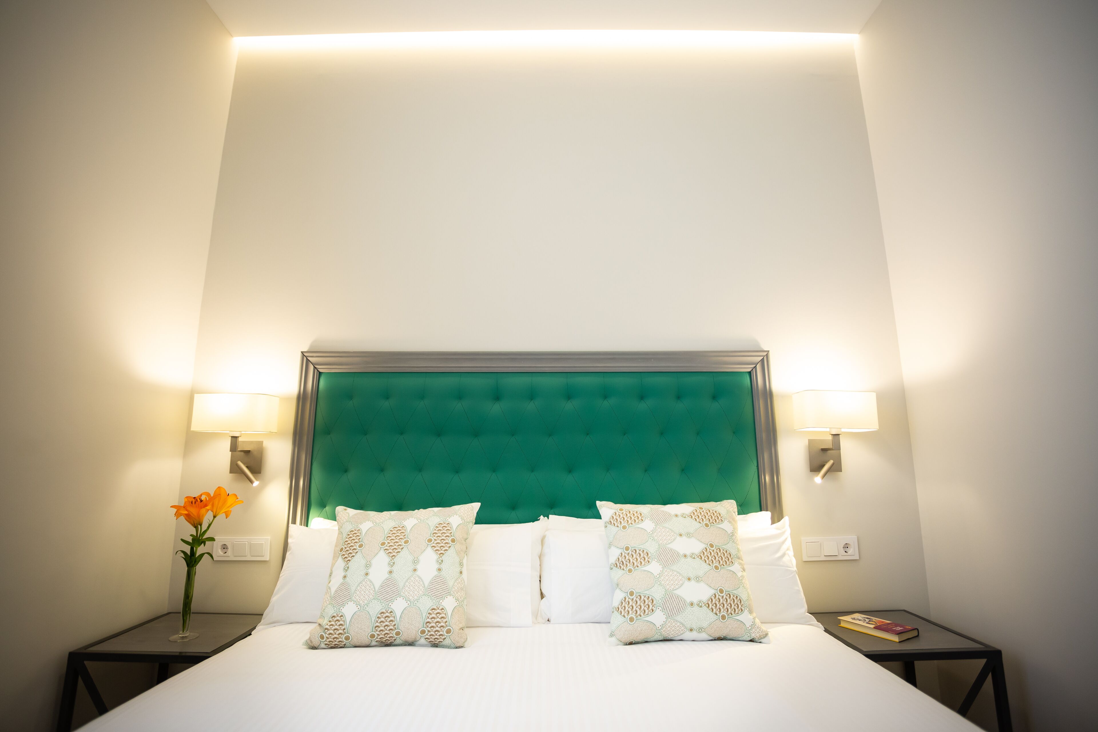 Una cama bien hecha con sábanas blancas y almohadas decorativas, flanqueada por dos mesitas de noche con lámparas y un florero.