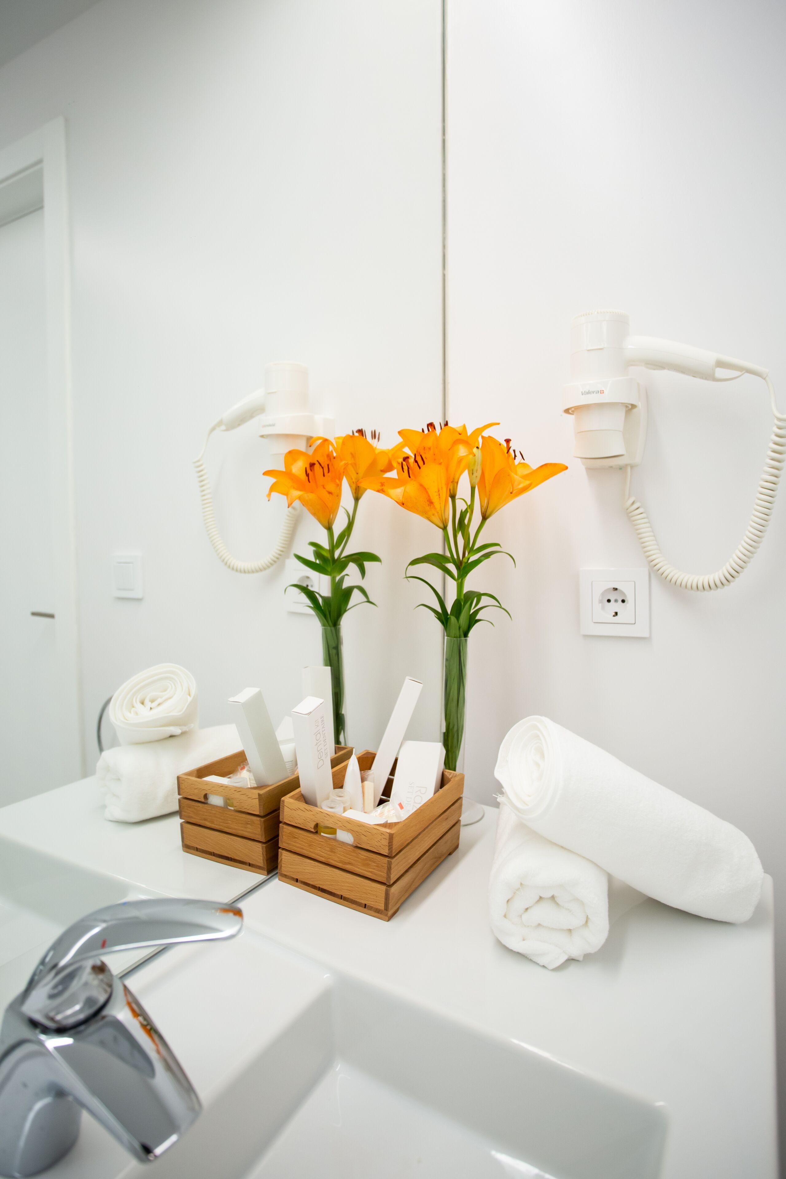 Une salle de bain avec un lavabo blanc, un miroir, un sèche-cheveux, une serviette et un vase avec des fleurs.