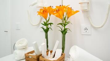 Une salle de bain avec un lavabo blanc, un miroir, un sèche-cheveux, une serviette et un vase avec des fleurs.