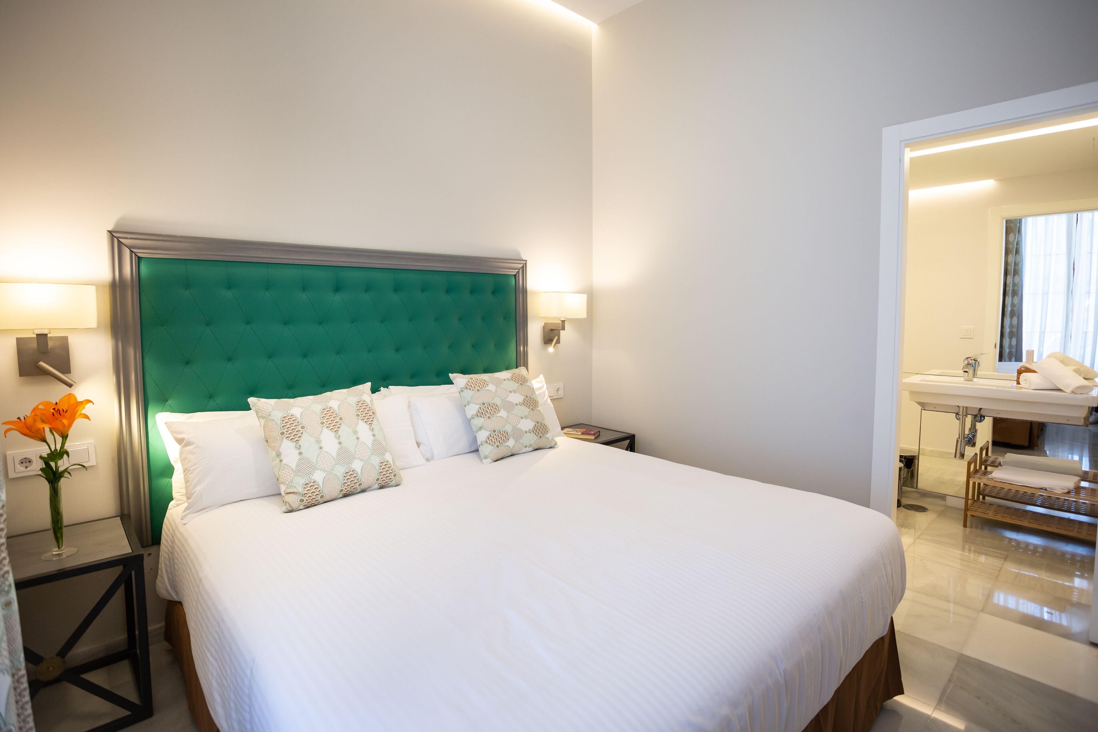 Una cama bien tendida con cabecera verde, ropa de cama blanca y almohadas estampadas. Hay una mesita de noche con una lámpara y un jarrón de flores. Se ve un baño al fondo con un lavamanos y un espejo.