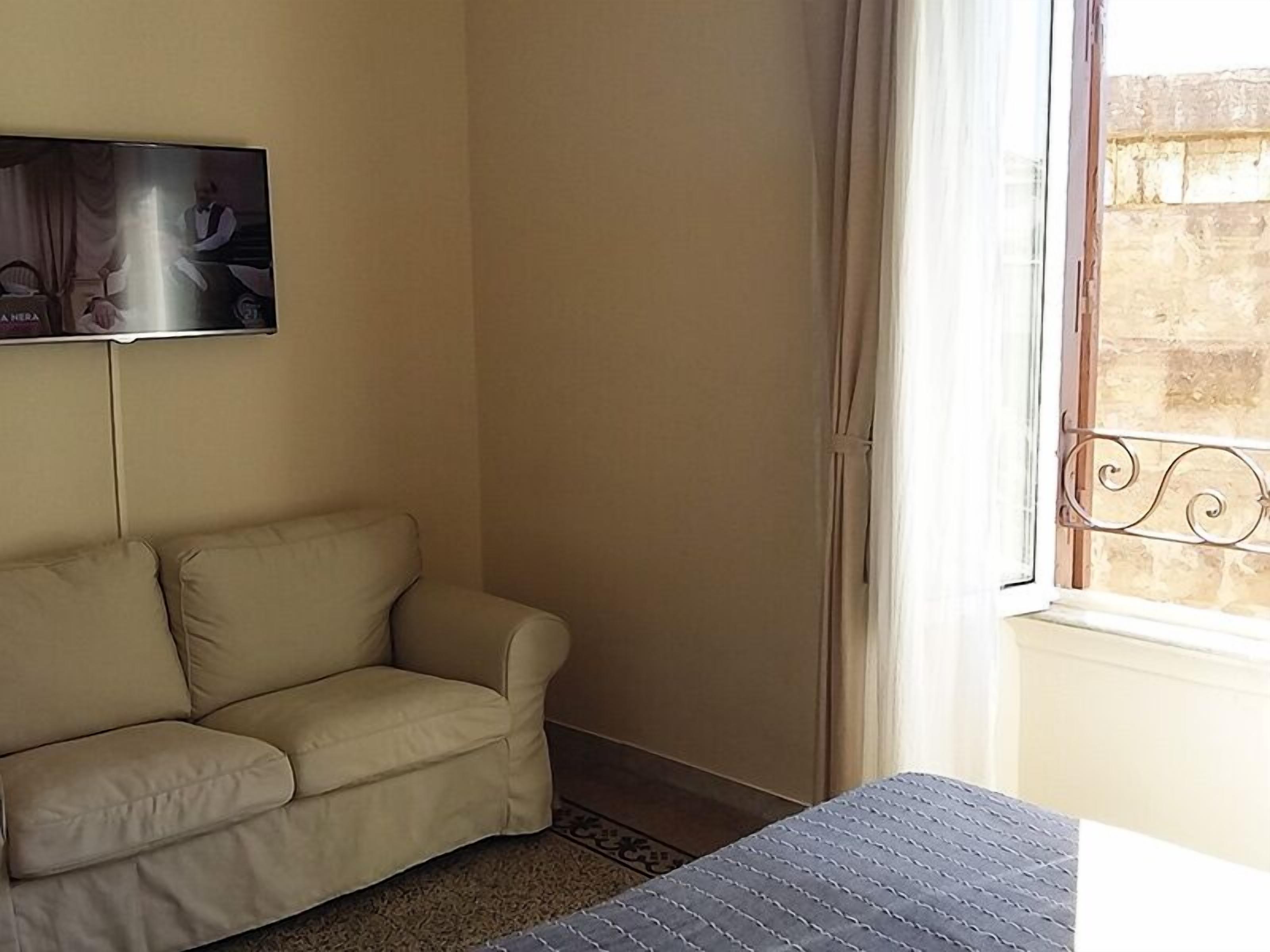 A room with a beige sofa, a television mounted on the wall, and a window with a metal railing.