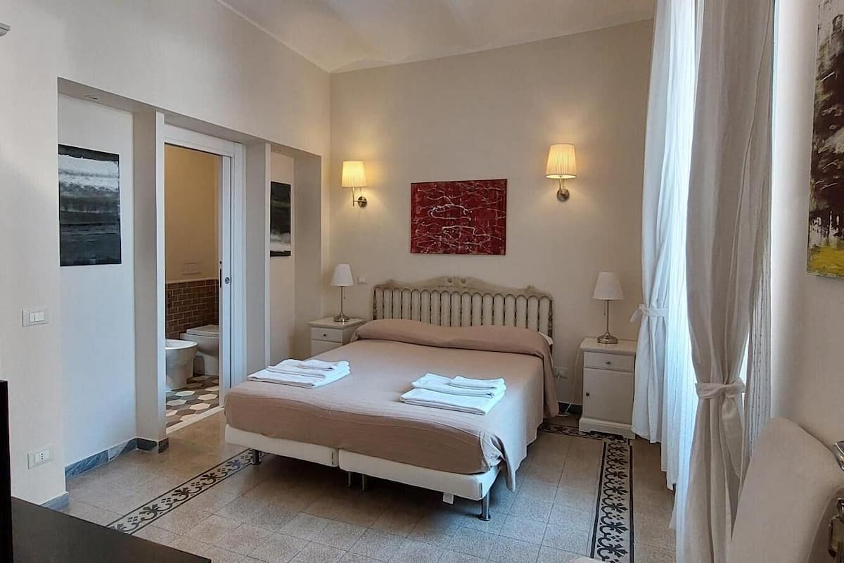 A hotel room with a bed, two bedside tables, a bathroom visible through an open door, and a painting on the wall.