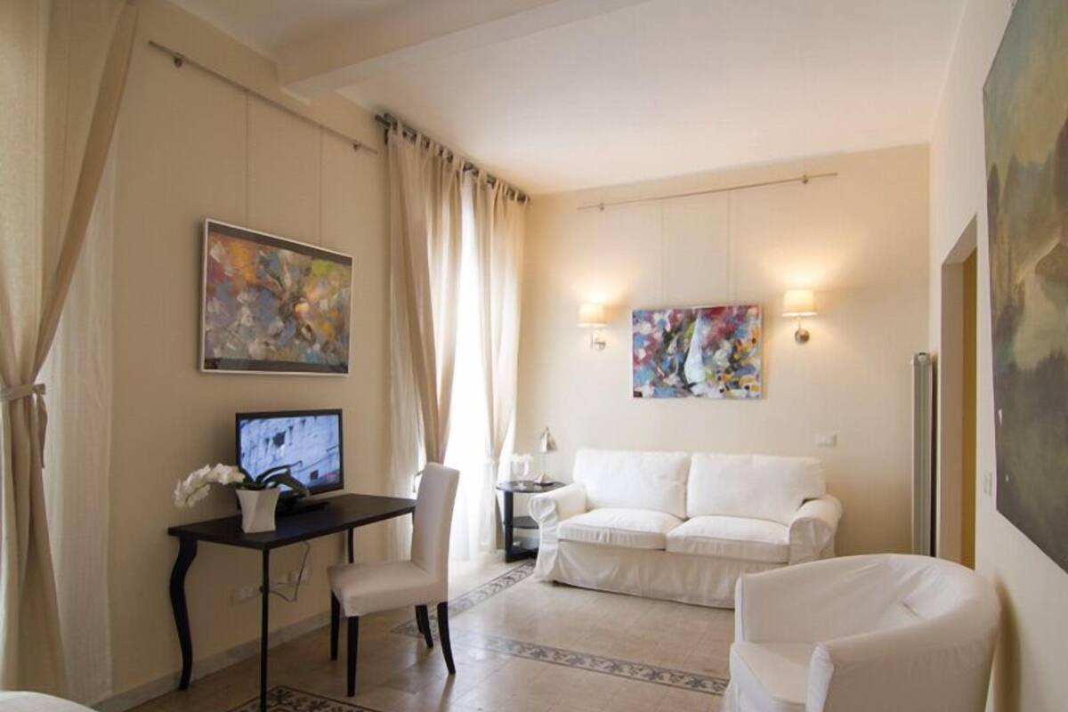 A hotel room with a white sofa, a desk with a computer, a chair, and two wall-mounted lamps.