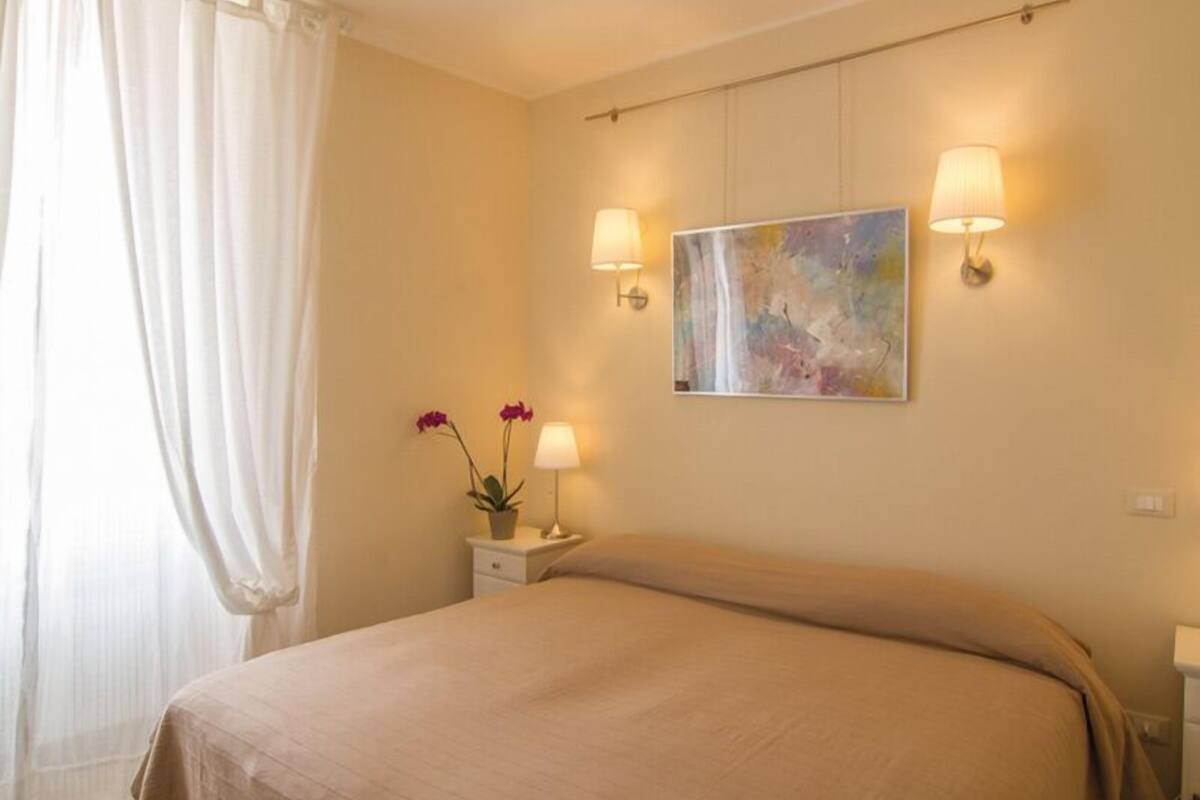 A bedroom with a bed, two bedside lamps, a nightstand, a painting, and a window with curtains.