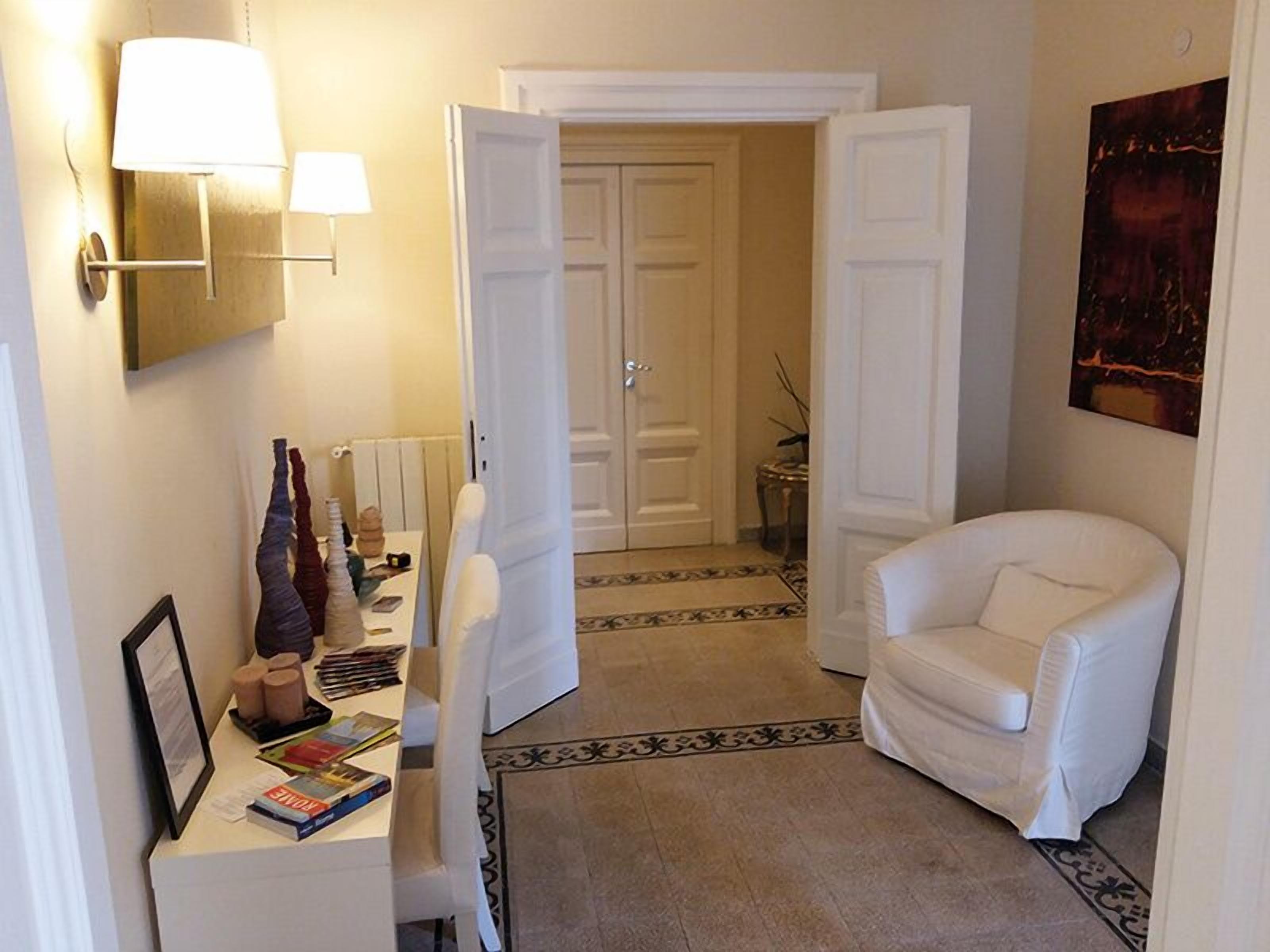 A hallway with a white chair, a table with books and decorative items, and two open doors leading to another room.