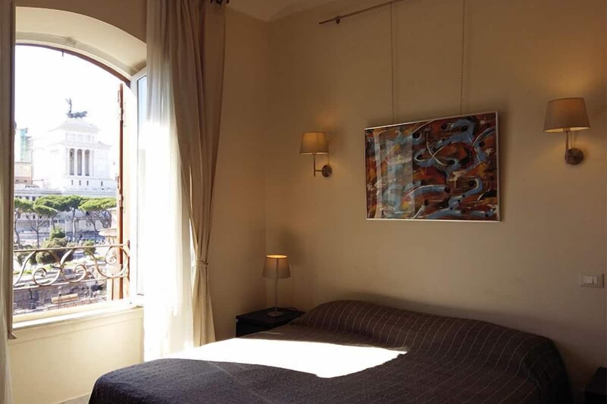 A hotel room with a bed, a painting, two wall sconces, a window with a view of a building, and a curtain.