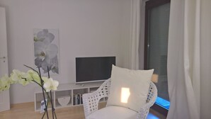 A living room with a white armchair, a flat-screen TV on a white stand, a white orchid plant, and a window with curtains. - Private apartment Angelika NEW (Lorch)
