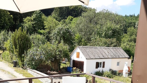 A wooden deck with a round table and white wicker chairs, overlooking a rural landscape with a white house and dense greenery. - Private apartment Angelika NEW (Lorch)