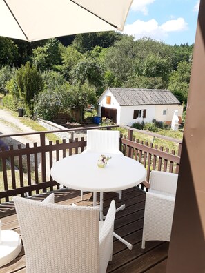 A wooden deck with a round table and white wicker chairs, overlooking a rural landscape with a white house and dense greenery. - Private apartment Angelika NEW (Lorch)