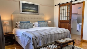 A neatly arranged bedroom with a large bed, bedside lamps, a wooden bench, and a view of the bathroom through a glass door. - Redbourne Hilldrop B&B (Cape Town)