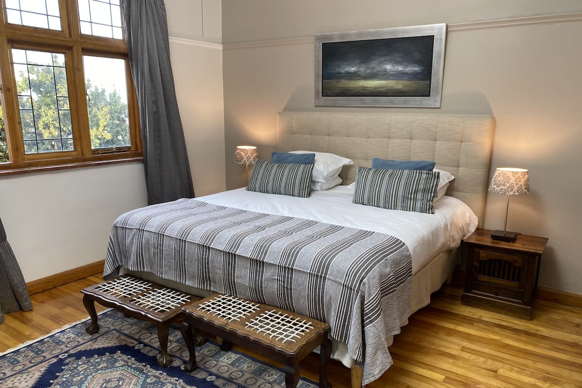 A bedroom with a large bed, two bedside tables with lamps, a bench, a patterned rug, and a window with curtains.