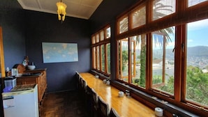 A room with a long wooden table, a refrigerator, and a large window with a view of greenery and buildings. - Redbourne Hilldrop B&B (Cape Town)