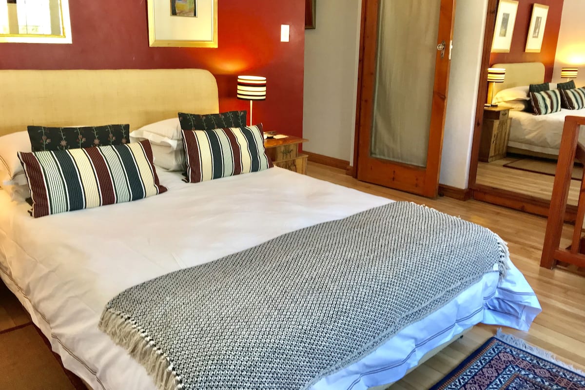 A hotel room with a large bed, a wooden headboard, a bedside table with a lamp, a mirror, and a door leading to another room.