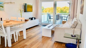 A modern living room with a dining area, a sofa, a coffee table, and a TV mounted on the wall. - Groyne 10 App No. 26 max 2 pers (+1 child 0-3 years) - 7/26 (Dierhagen)