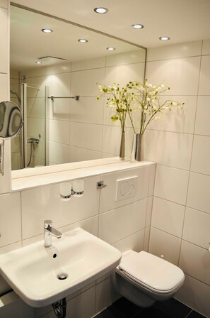 A modern bathroom with a white sink, toilet, and a mirror above the sink. There are two vases with flowers on the sink counter.