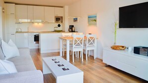 A modern kitchen with white cabinets, a dining area with white chairs, a flat-screen TV mounted on the wall, and a coffee maker. - Groyne 10 App No. 26 max 2 pers (+1 child 0-3 years) - 7/26 (Dierhagen)
