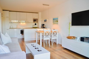A modern kitchen with white cabinets, a dining area with white chairs, a flat-screen TV mounted on the wall, and a coffee maker.
