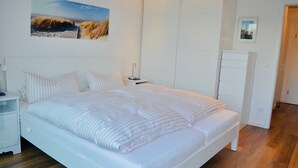 A bedroom with a large bed, a nightstand, a dresser, and a wall-mounted picture. - Groyne 10 App No. 26 max 2 pers (+1 child 0-3 years) - 7/26 (Dierhagen)