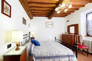 A bedroom with a wooden ceiling, a bed with a patterned duvet, a nightstand with a lamp, a dresser, and a chair.